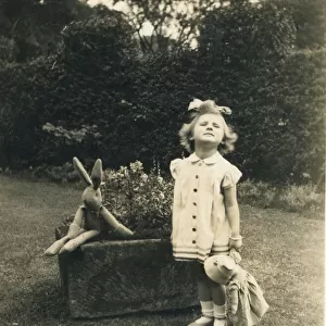 A little girl stands to attention and grimaces for the camera in a garden holding a doll