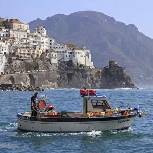Fisherman in fishing boat heads out to sea from Amalfi harbour, with view towards Amalfi town