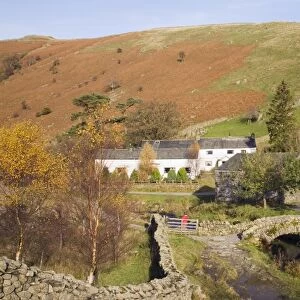 Old stone packhorse bridge over Watendlath Beck with dry stone wall and farm buildings in picturesque village in autumn, Watendlath, Lake District National Park, Cumbria, England, United