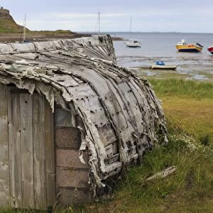 Upturned weathered boat hut with Lindisfarne Castle and fishing boats at low tide