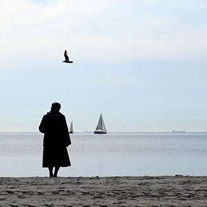A woman, wrapped up against the cold wind, stands on the beach as she watches a swan