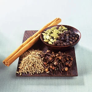 Cardamom pods, cinnamon sticks, cloves, cumin seeds, and black peppercorns arranged in wooden bowl and on wooden board