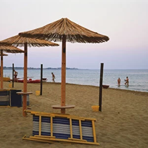 Italy, Sicily, Southeast Sicily, Noto, folded deckchairs propped against bamboo umbrellas on almost deserted beach, with some people paddling in sea, sunset, blurred motion