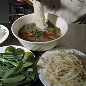 Man using chopsticks to lift rice noodles from bowl of Pho meat soup, plates of bean spouts and green leaves on table, Ho Chi Minh City