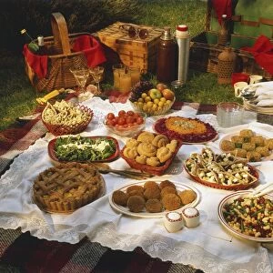 Picnic display, baskets, thermos flasks, bottles, glasses, bowls and dishes with salads, breaded balls, burgers, quiches, tomatoes, fruit, arranged on white lace blanket, checked picnic rug underneath, grass background
