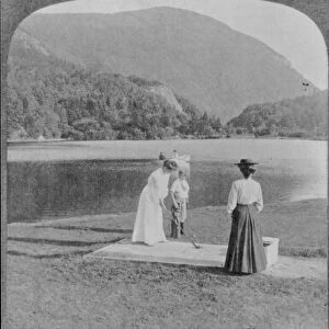 Woman and boy with golf clubs in foreground, 2 people in boat on lake, mountain in background