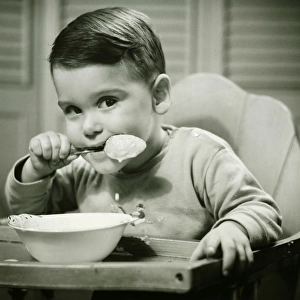 Boy (4-5) sitting in high chair, eating porridge, (B&W), portrait