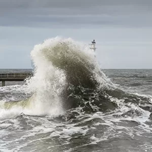 Large Wave Crashing At The Coast With A Lighthouse At The End Of A Pier; Amble, Northumberland, England