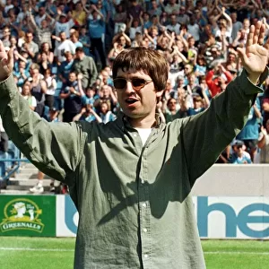 Liam Gallagher arms raised, stands on a pitch at Maine Road during the Premiership match