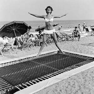 A model wearing bikini swimwear bounces on a trampoline on a beach in the French Riviera