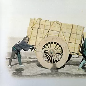Portrait of three japanese carters. Two of the three men are at the head of a wagon and are pulling it while the third is pushing from behind. The wagon is full of large bundles