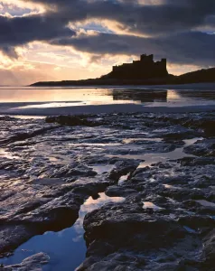 Prints of BAMBURGH CASTLE, Northumberland. The coastal castle viewed from the beach, silhouetted against a setting sun Explore England - Iconic Landmarks and Places to Visit: Bamburgh Castle Print at Sunset