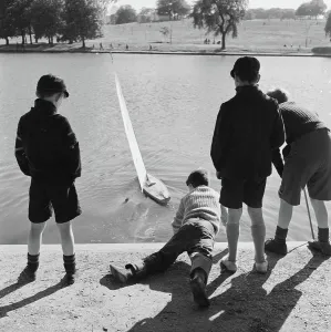 Prints of HIGHGATE PONDS, Hampstead Heath, Greater London. Three boys standing watching a model yacht being launched on one of the Highgate Ponds by a boy lying on his front reaching over the water. John Gay Iconic Photographers of England - Historic Image Collections: Print of Highgate Ponds, Hampstead Heath