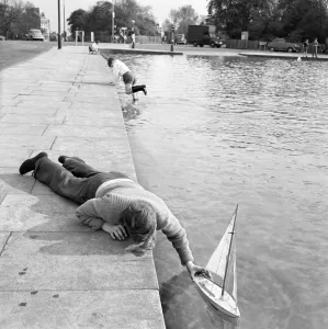 Prints of WHITESTONE POND, Hampstead Heath, London. View of a boy lying down on the edge of the pond with his hand holding a model yacht on the water Iconic Photographers of England - Historic Image Collections: Whitestone Pond Print, Hampstead Heath