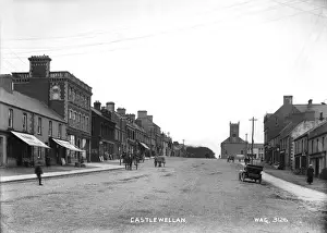 Prints of Castlewellan - a view up a hill with a wide road, with shop fronts both sides and motor vehicles and horse vehicles and people. At the top of the hill is a Church Castlewellan Prints: Castlewellan Castlewellan Print: Early 20th Century Scene