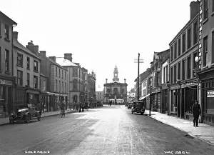 Prints of Coleraine - a busy street scene with shop fronts, motor cars, people and Clock Tower with motor bus outside. (Location: Northern Ireland; County Londonderry; Coleraine). Date: circa early 1900s Derry Prints: Coleraine Print: Early 20th Century Street Scene