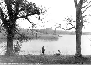 Prints of Downpatrick from Inch Abbey - a view of the Cathedral in the distance and in the foreground sits a girl and a boy is standing fishing, framed by two trees Down Prints: Early 20th Century Print of Downpatrick from Inch Abbey