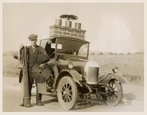 Prints of A chimney sweep stands with his equipment beside his car which has a model of a chimney on the roof with a broom sticking out of the top, to advertise his trade S Prints: Mobile Chimney Sweep Print