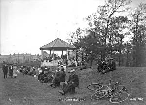 Prints of The Park, Bangor - a view of a lot of people sitting on benches and grassy mound beside a Bandstand in Ward Park. In the foreground are bicycles and in the distance buildings Down Prints: Early 20th Century Print of Ward Park, Bangor