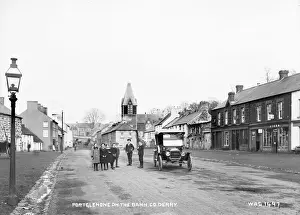 Prints of Portglenone on the Bann, Co. Derry - a street scene with a motor car and children in the foreground and horse vehicles in the distance. There are some shop fronts and thatched buildings County Londonderry Prints: Early 20th Century Print of Portglenone Street Scene