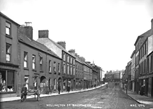 Prints of Wellington Street. Ballymena - a street scene with people and boy and hand cart. To the distance can be seen a horse vehicle and motor car. (Location: Northern Ireland: County Antrim) County Antrim Prints: Early 20th Century Ballymena Street Scene Print
