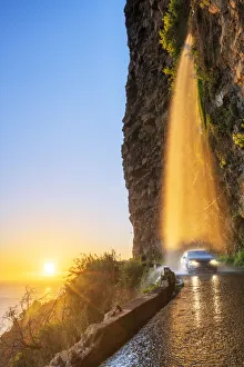 Car passing under Anjos waterfall on slippery coastal road at sunset, Ponta do Sol Car passing under Anjos waterfall on slippery coastal road at sunset, Ponta do Sol