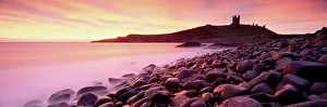 An imposing silhouette of Dunstanburgh Castle against a magnificent sky at sunrise with a beach of basalt boulders in An imposing silhouette of Dunstanburgh Castle against a magnificent sky at sunrise with a beach of basalt boulders in