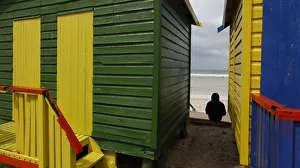 A spectator sits between beach huts as surfers prepare to take to the water in an A spectator sits between beach huts as surfers prepare to take to the water in an