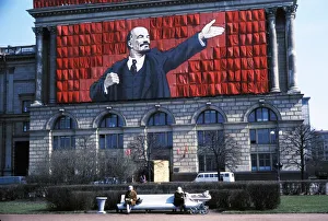 A banner of lenin on the side of a building overlooking two elderly women sitting on a park bench in leningrad, ussr A banner of lenin on the side of a building overlooking two elderly women sitting on a park bench in leningrad, ussr