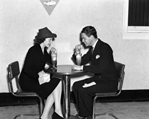 Couple In Luncheonette Both Are Sitting At A Table Wearing Dark Suits & Eating An Couple In Luncheonette Both Are Sitting At A Table Wearing Dark Suits & Eating An