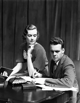 Couple Working On Budget At Desk Woman Sitting On Arm Of Chair Man With Pencil Writing On Couple Working On Budget At Desk Woman Sitting On Arm Of Chair Man With Pencil Writing On