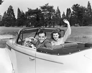 Teenage couple in convertible car smile and wave to viewer Teenage couple in convertible car smile and wave to viewer