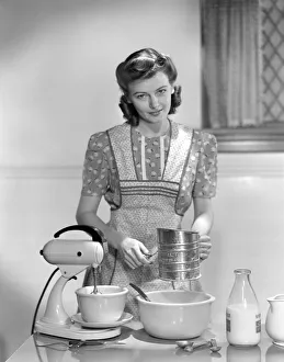 Woman In A Apron Over A Cotton Print Dress Sifting Flour In A Bowl Between A Mixer & A Woman In A Apron Over A Cotton Print Dress Sifting Flour In A Bowl Between A Mixer & A