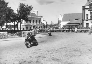 Competitors passing Ramsay Town Square at speed. Number 69 is W. J. Evans on AJS and number 90 is C Competitors passing Ramsay Town Square at speed. Number 69 is W. J. Evans on AJS and number 90 is C
