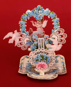 A pop-up card a winged cherub playing a flute under a heart shaped arch of forget-me-nots A pop-up card a winged cherub playing a flute under a heart shaped arch of forget-me-nots