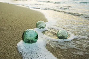 Three Glass Fishing Floats Roll On The Sandy Shoreline With Ripples Of Water And Seafoam Three Glass Fishing Floats Roll On The Sandy Shoreline With Ripples Of Water And Seafoam