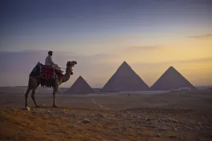 A Lone Camel And Rider Stand In Front Of The Setting Sun With The Great Pyramids Behind Them; Giza, Egypt A Lone Camel And Rider Stand In Front Of The Setting Sun With The Great Pyramids Behind Them; Giza, Egypt