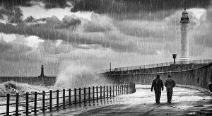 Two People Walking Along The Water's Edge In The Rain With Waves Crashing Into The Shore And A Lighthouse In The Two People Walking Along The Water's Edge In The Rain With Waves Crashing Into The Shore And A Lighthouse In The