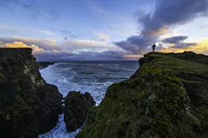 Person Standing On A Cliff Face While The Atlantic Ocean Pounds The Shores Beneath Them; Iceland Person Standing On A Cliff Face While The Atlantic Ocean Pounds The Shores Beneath Them; Iceland
