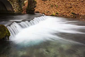 Water Cascading Over A Ledge In A River With The Riverbank Covered With Autumn Coloured Water Cascading Over A Ledge In A River With The Riverbank Covered With Autumn Coloured