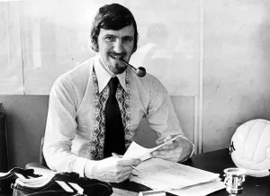 Jimmy Hill sitting at his desk smoking a pipe, dressed in stylish 1970s clothing Jimmy Hill sitting at his desk smoking a pipe, dressed in stylish 1970s clothing