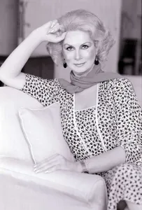 Model April Ashley sitting on a sofa wearing a print dress with one hand at her head May Model April Ashley sitting on a sofa wearing a print dress with one hand at her head May