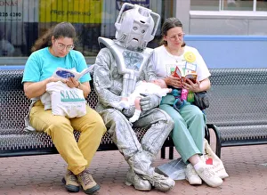Person in a robot outfit sits down on a bench next to two women shoppers reading their Person in a robot outfit sits down on a bench next to two women shoppers reading their