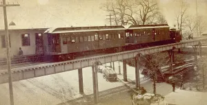 View of an elevated railway structure with train that has pulled into the station, in New York city. In the street below some freight wagons are parked