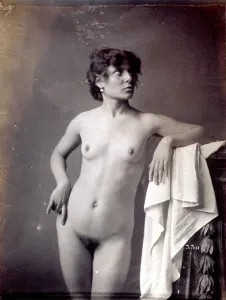 Young model shown, nude, in the studio. She is posing with her left arm resting on a pedestal covered by a light cloth and with her head facing to one side