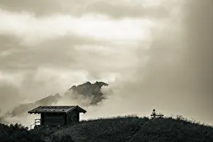 Female Hiker Prints: Mountain hut with bench and climber and dramatic clouds in the background