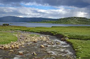 Cloudy Sky With Mountains Prints: A small river flows through green fields under a cloudy sky with mountains in the background